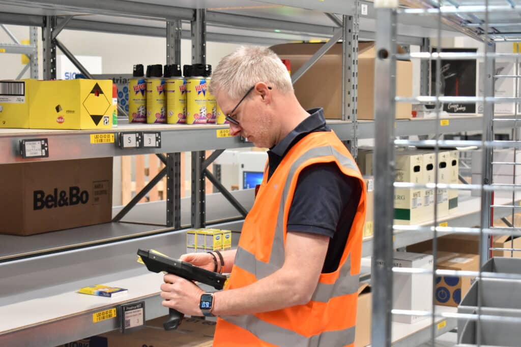 Magazijnmedewerker met oranje veiligheidsvest scant producten op rekken met een handheld scanner in het testcentrum van Fabri