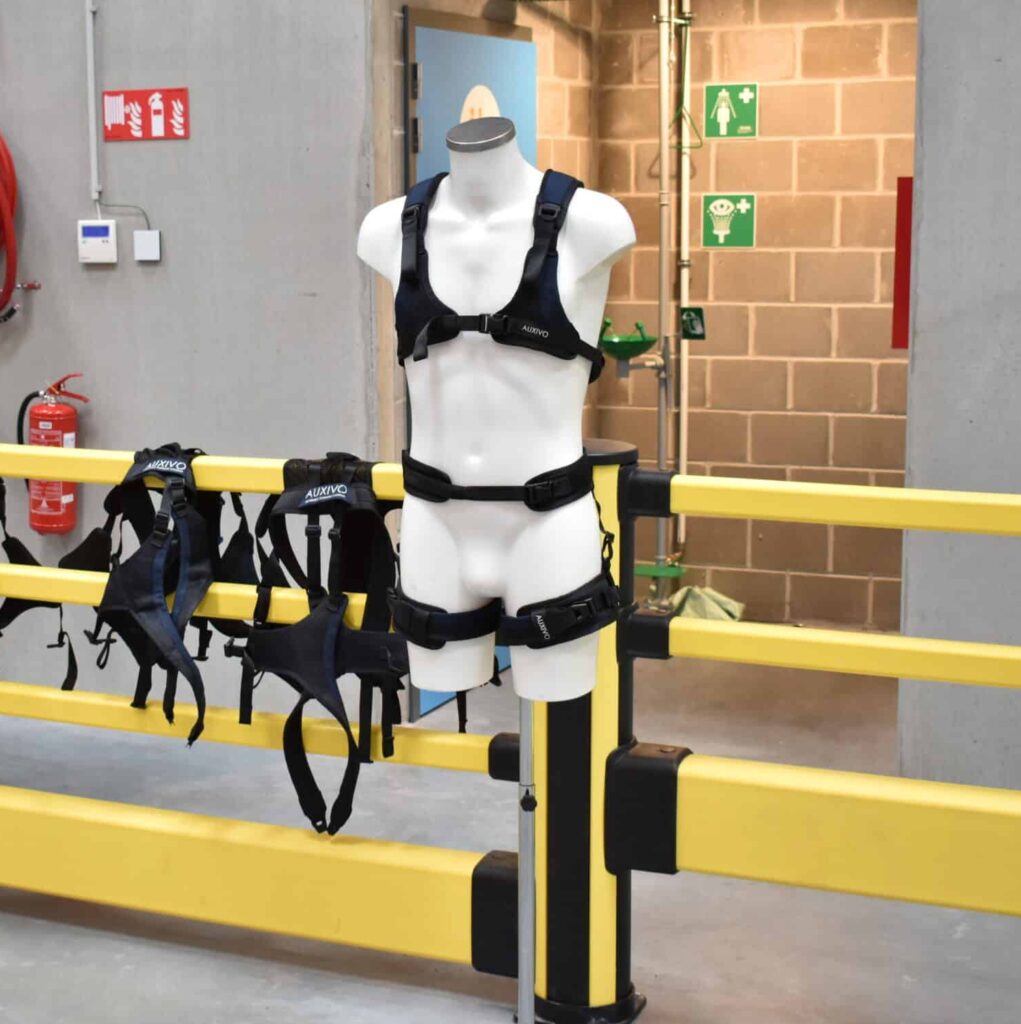 Auxivo passief exoskelet op mannequin in testcentrum Fabriek Logistiek, met meerdere harnasvarianten op gele veiligheidshekke